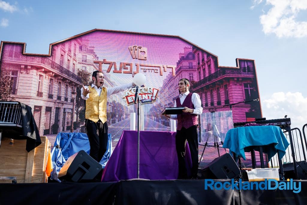 Mechy and Shloimy on an outdoor stage in gold and maroon vests, their name lit up on the LED wall behind them